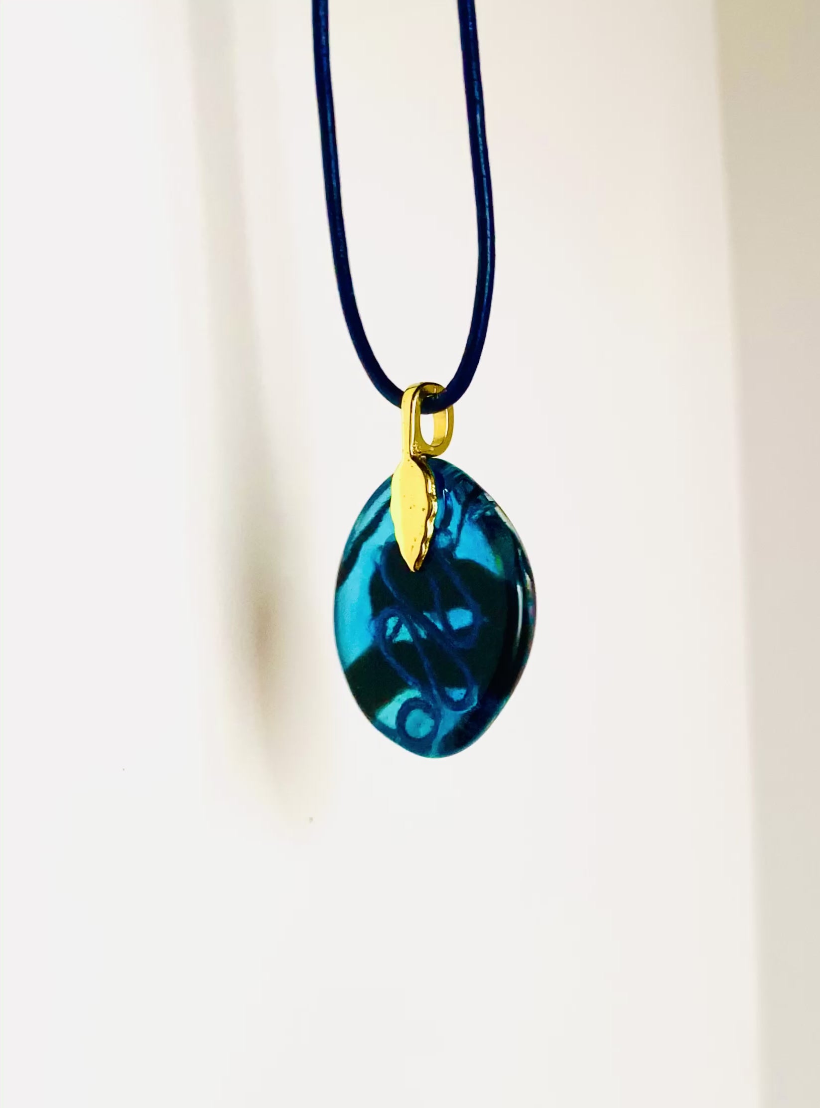 Video of fused glass pendant on a blue leather cord. Transparent blue glass with layers of red, rust, blue, green and purple dichroic glass and a copper squiggle design embedded in the glass. By 3D Glass Designs 