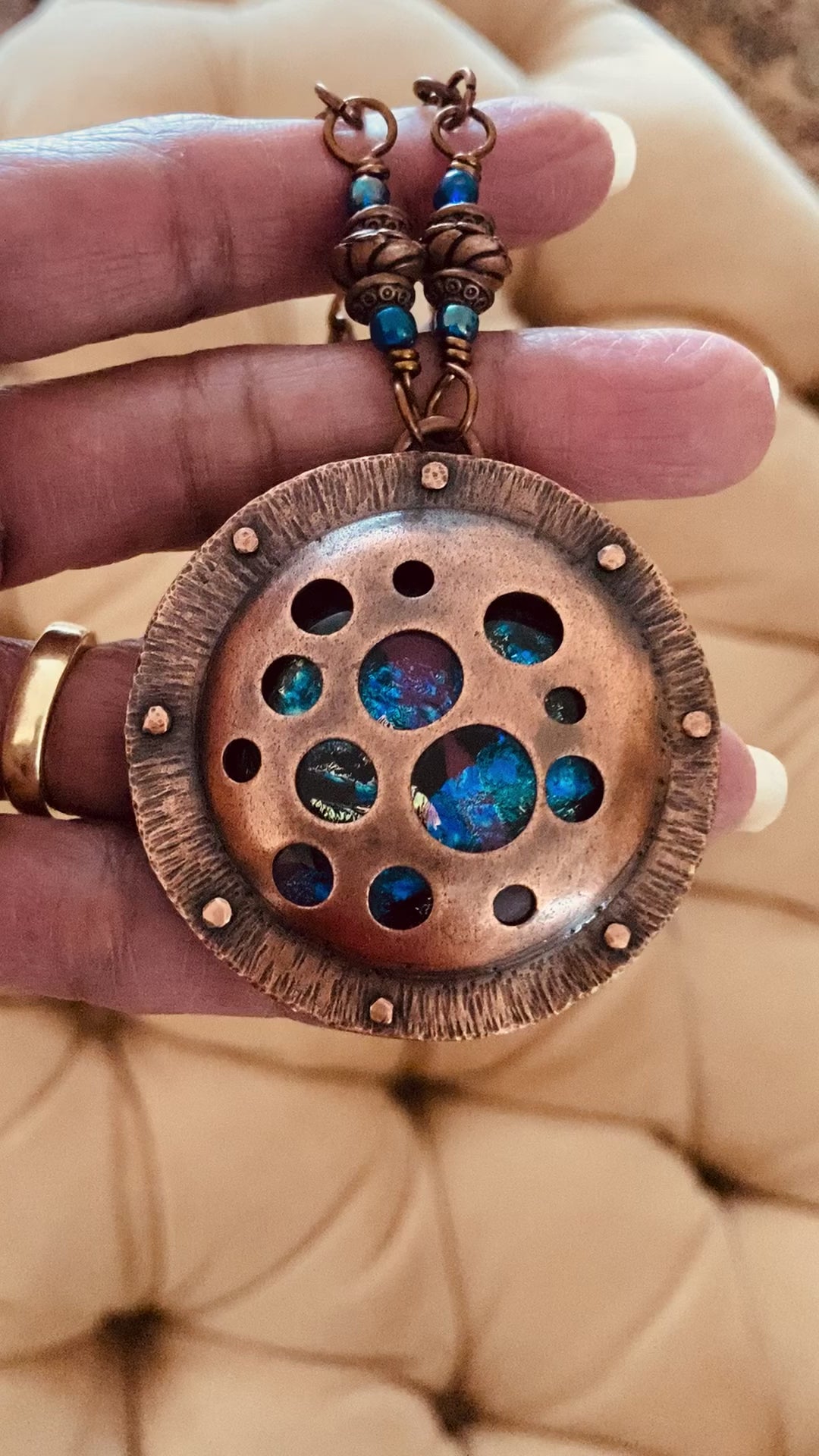 Video of Round copper pendant with various sized holes revealing pink and blue fused glass beneath, sandwiched between two layers of copper. This unique artisan pendant is hung from a beaded copper chain, by 3D Glass Designs.