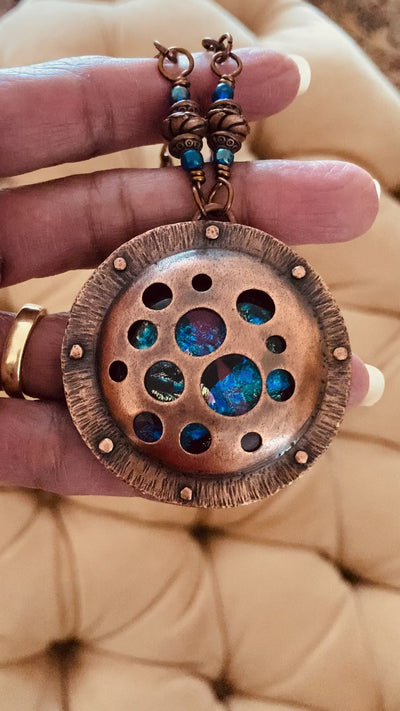 Video of Round copper pendant with various sized holes revealing pink and blue fused glass beneath, sandwiched between two layers of copper. This unique artisan pendant is hung from a beaded copper chain, by 3D Glass Designs.