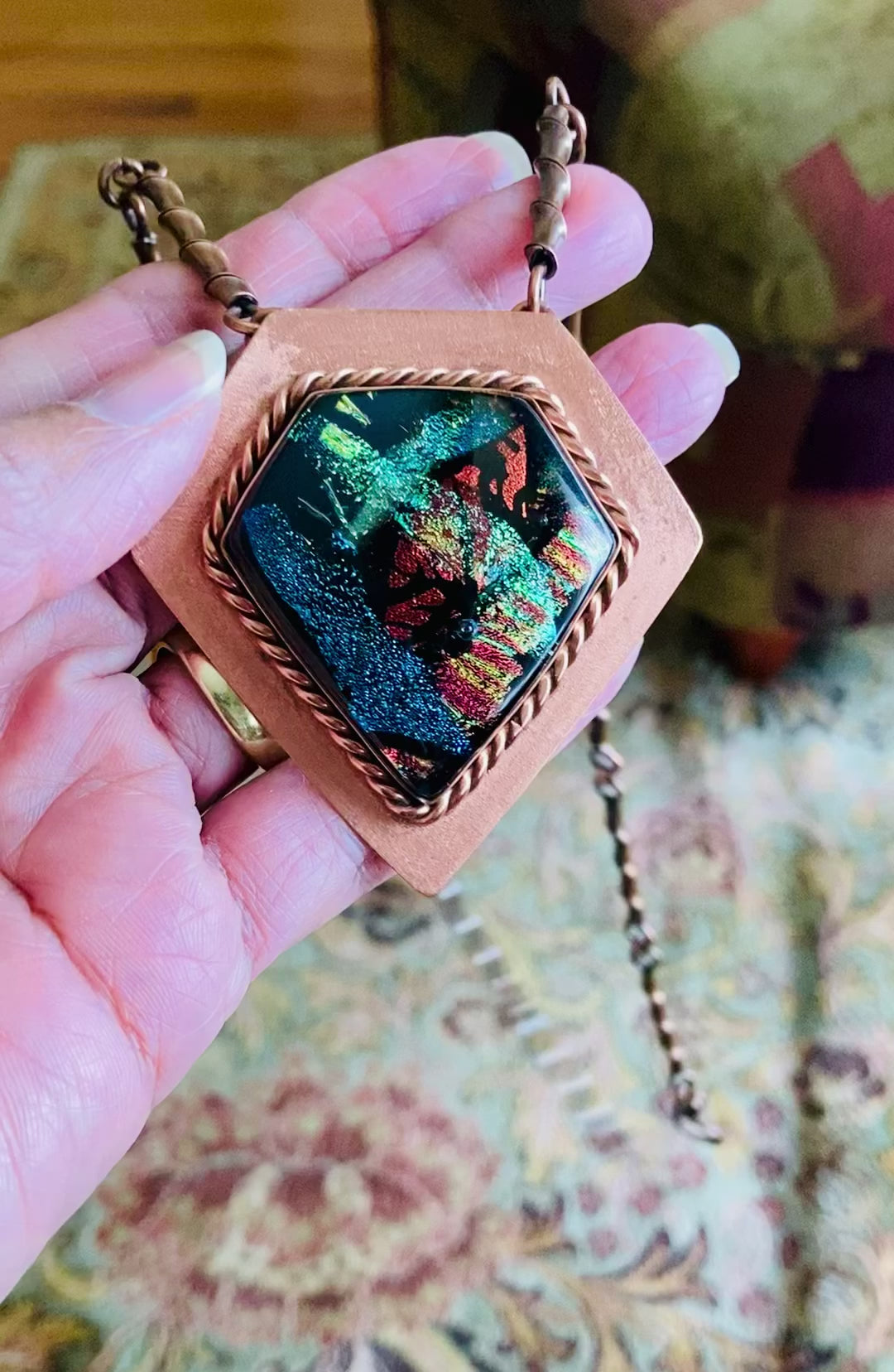 Video of model's hand showing the details of fused glass and copper pendant necklace by 3D Glass Designs.  Dichroic glass in blue, green and rust hues set in a handcrafted diamond-shaped shield copper pendant with an elegant rope detail surrounding the bezel.  The handmade copper necklace consists of twisted copper links that resembles bamboo and a handmade clasp.