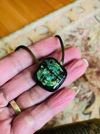 Hand holding green glass pendant on an olive-green satin cord against a sage green floral rug, by 3D Glass Designs.